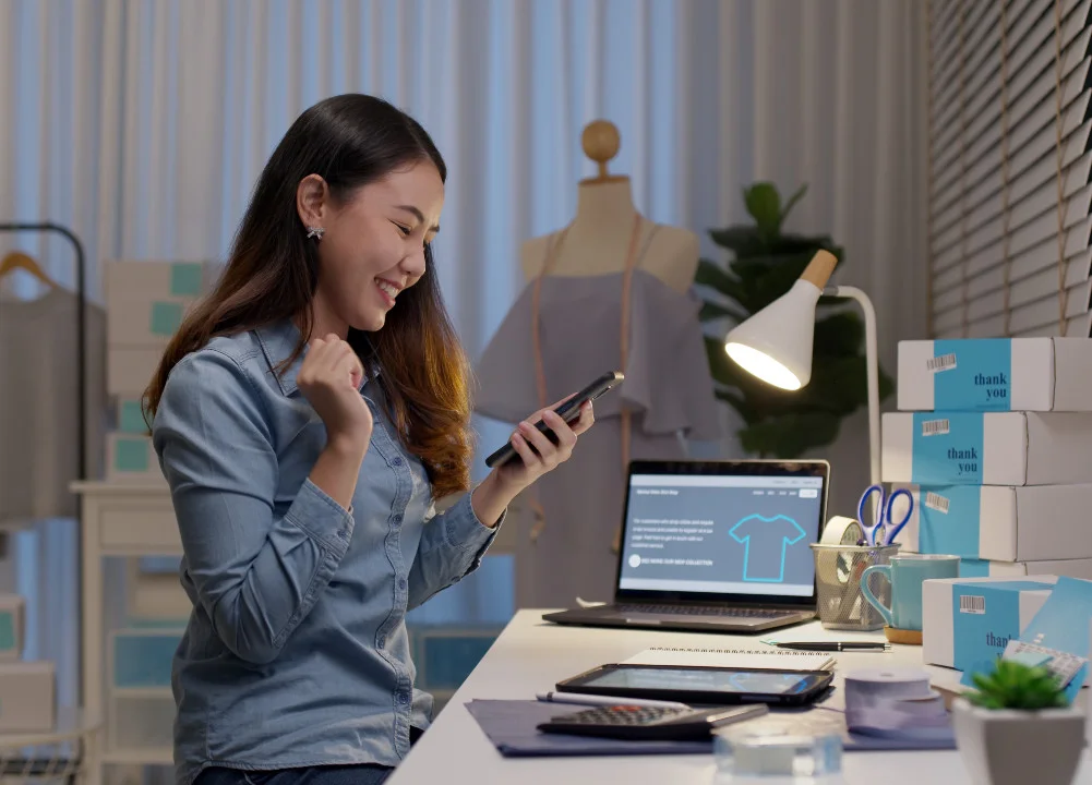 Female business owner sitting at a desk with her packages, looking at her phone. A laptop open to a product page, iPad, calculator and supplies are on her desk. In the background are more business supplies.